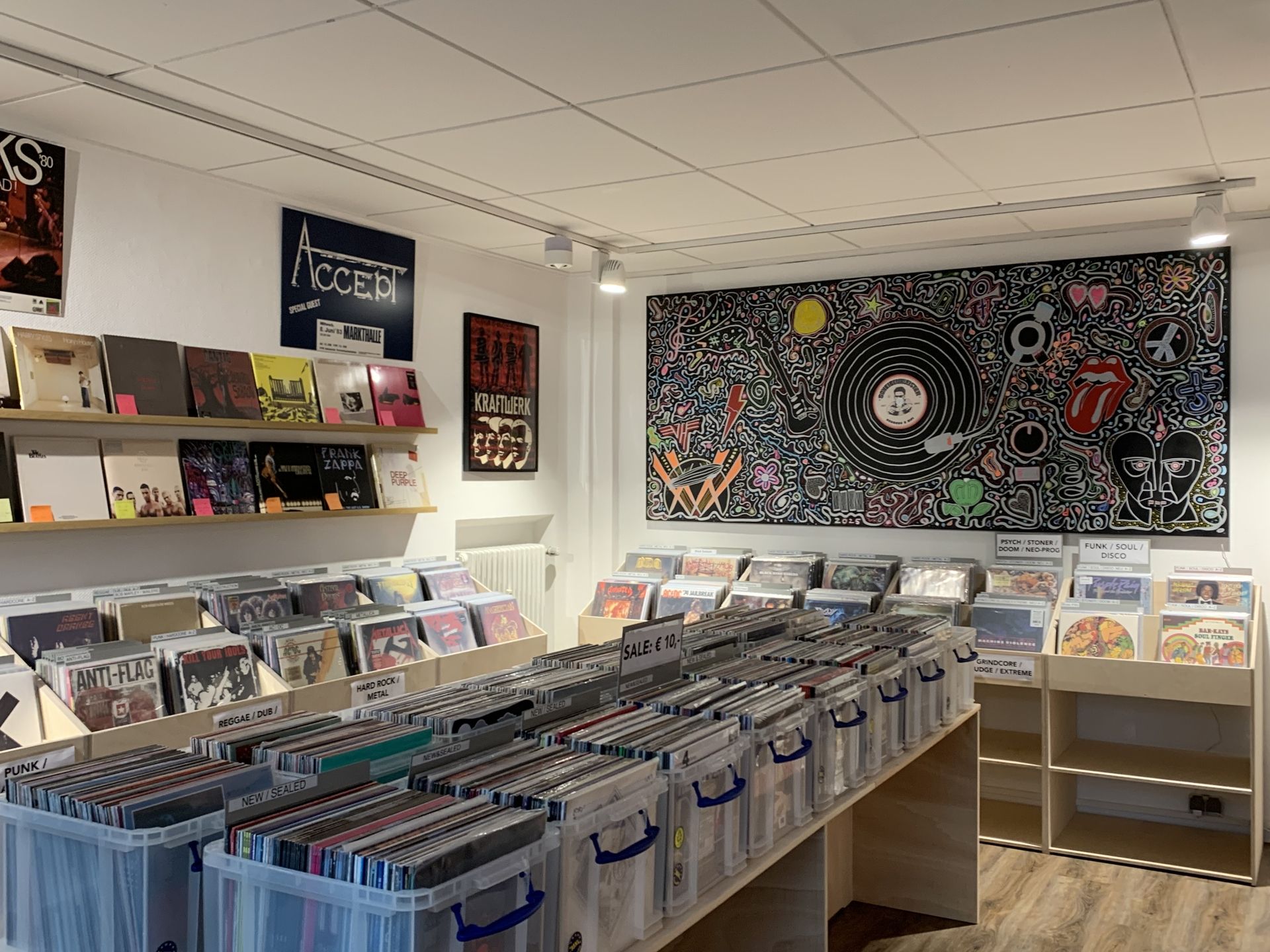 Innenansicht eines Plattenladens mit Regalen voller Schallplatten und einer Wand mit einem großen, bunten Wandbild mit Musik- und Popkulturmotiven