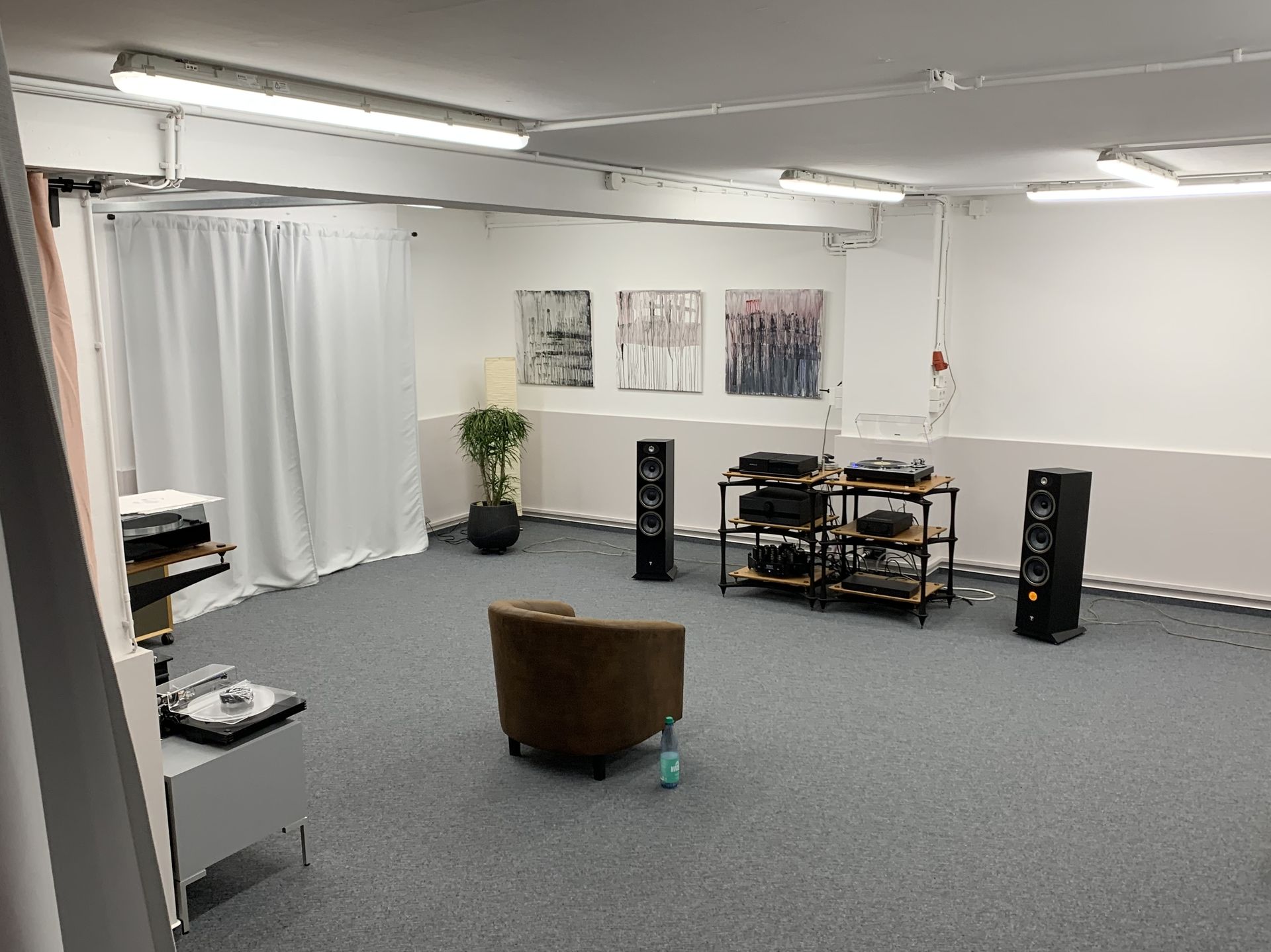 Leerer Hörraum mit einem braunen Sessel, zwei Lautsprechern, einem Regal mit Audioequipment und abstrakten Bildern an der Wand